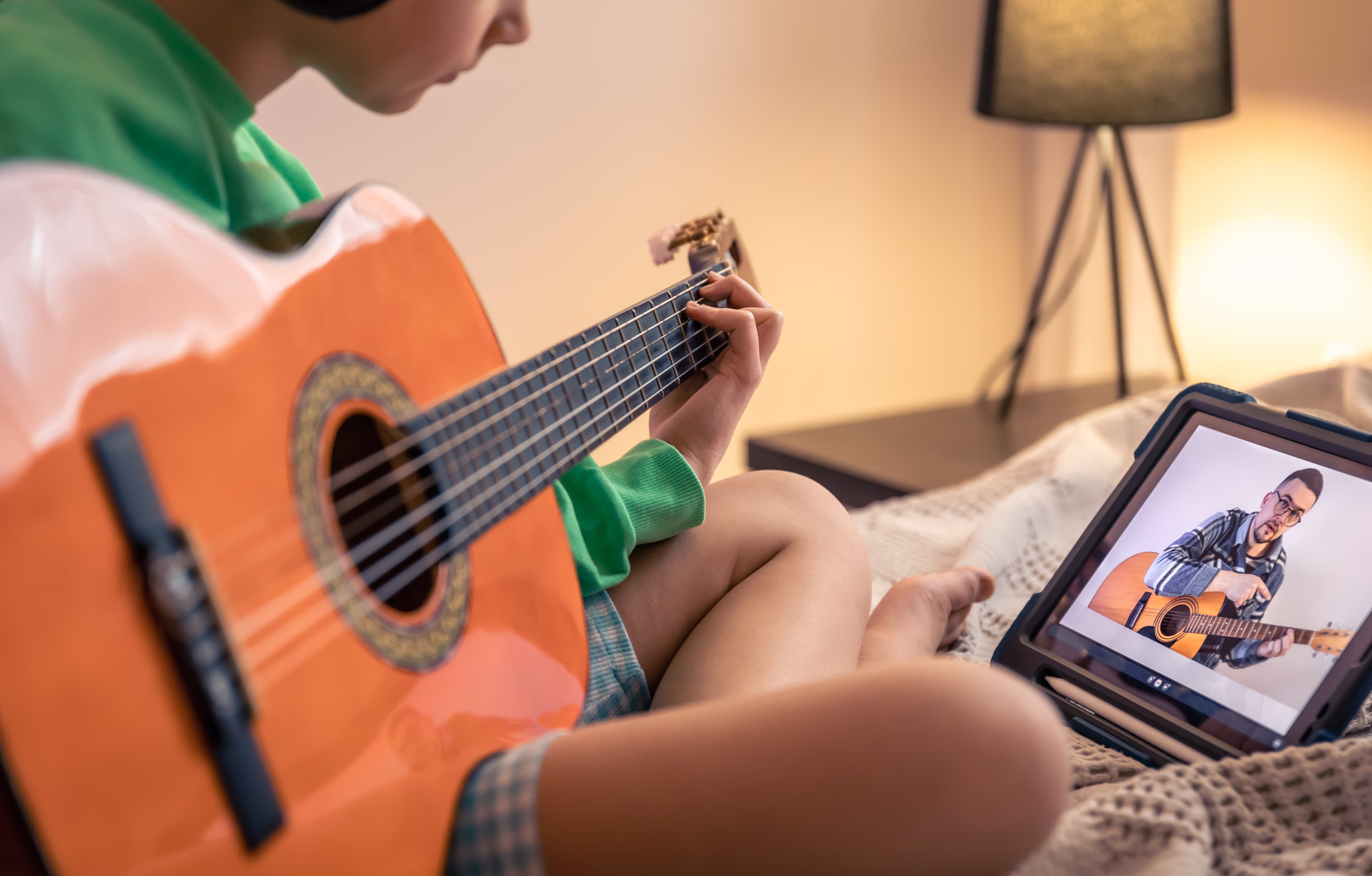 Fotografia fotorealist de um garoto sentado na cama com violão no colo assistindo aula de violão no tablet.